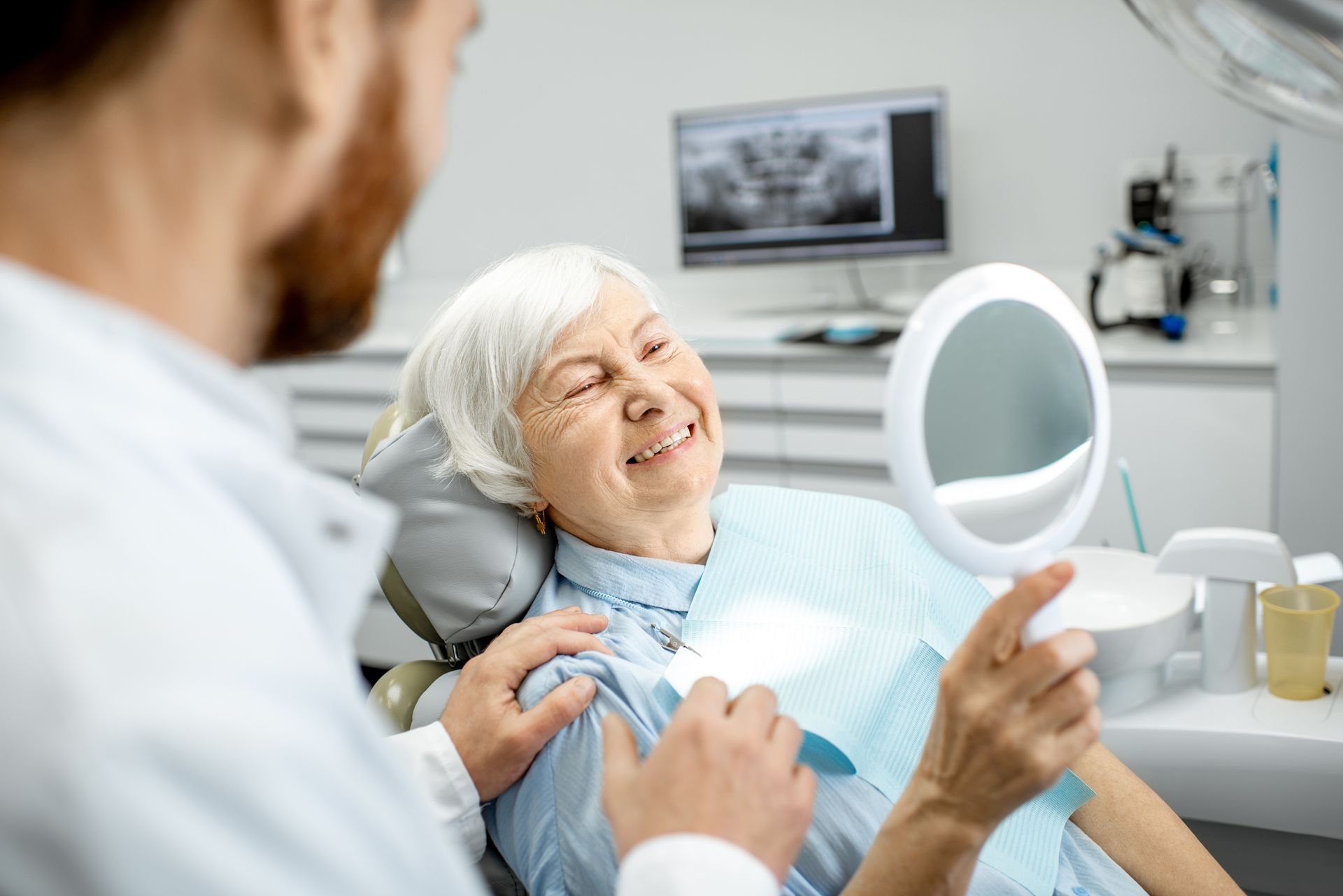 An elderly woman enjoys her beautiful smile, looking in the mirror, after local denture services.