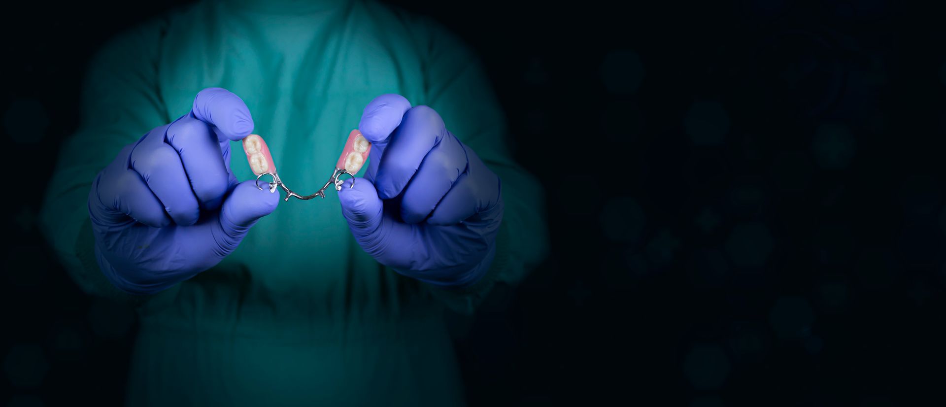A person in scrubs and gloves holding a dental bridge against a dark background.