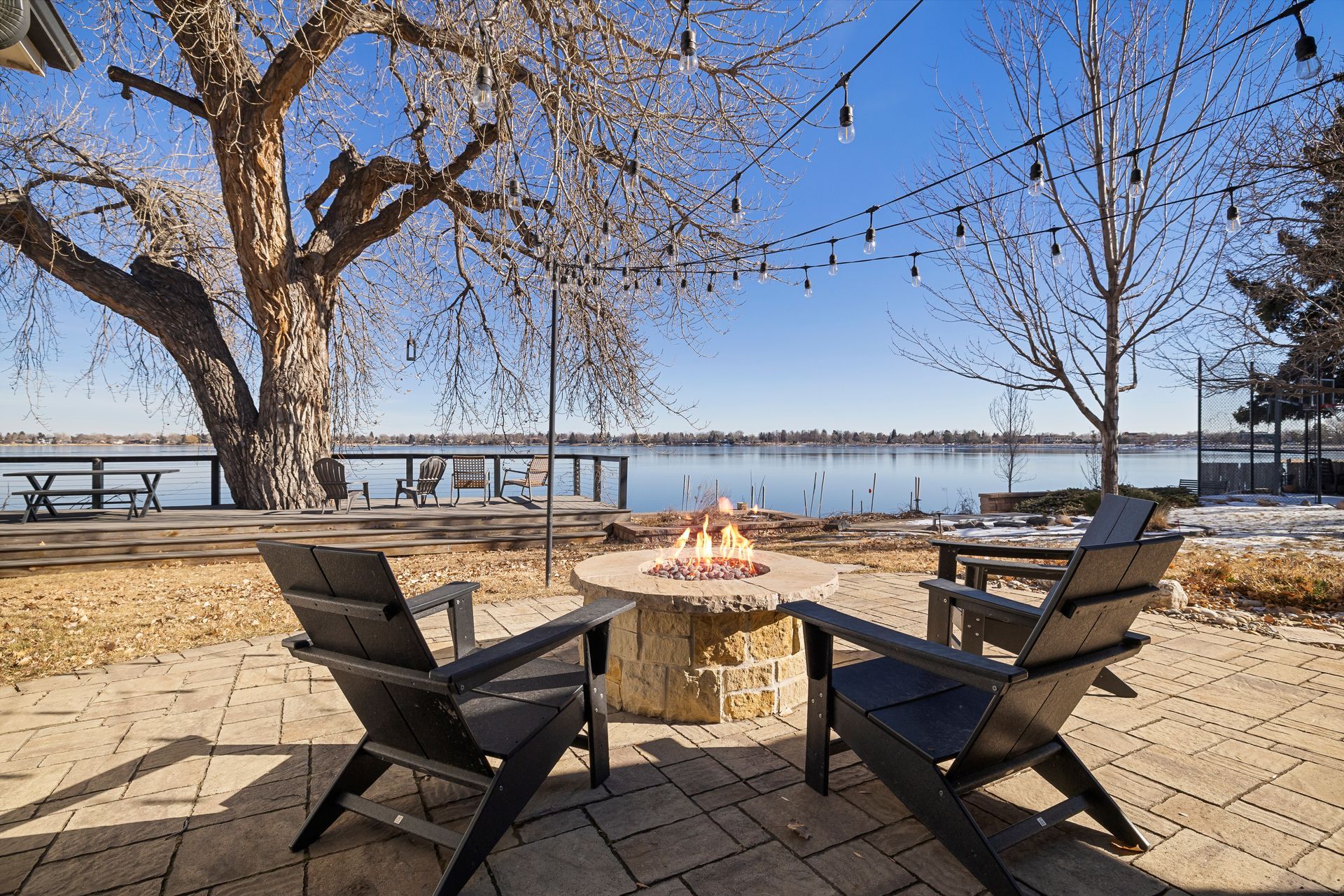 Two black chairs face a fire pit on a patio overlooking a lake, string lights overhead.