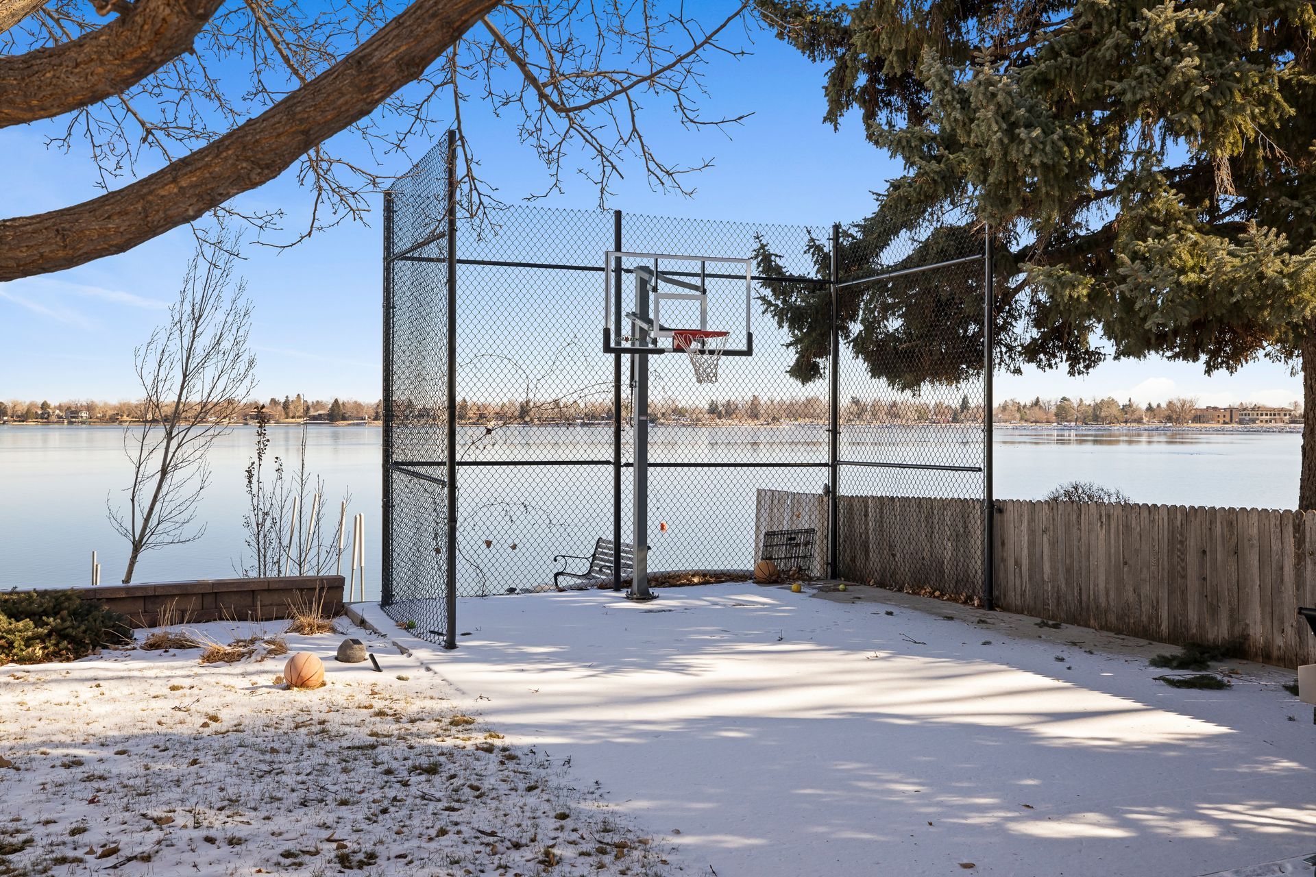 Basketball hoop on a snowy court beside a lake, enclosed by a net fence.