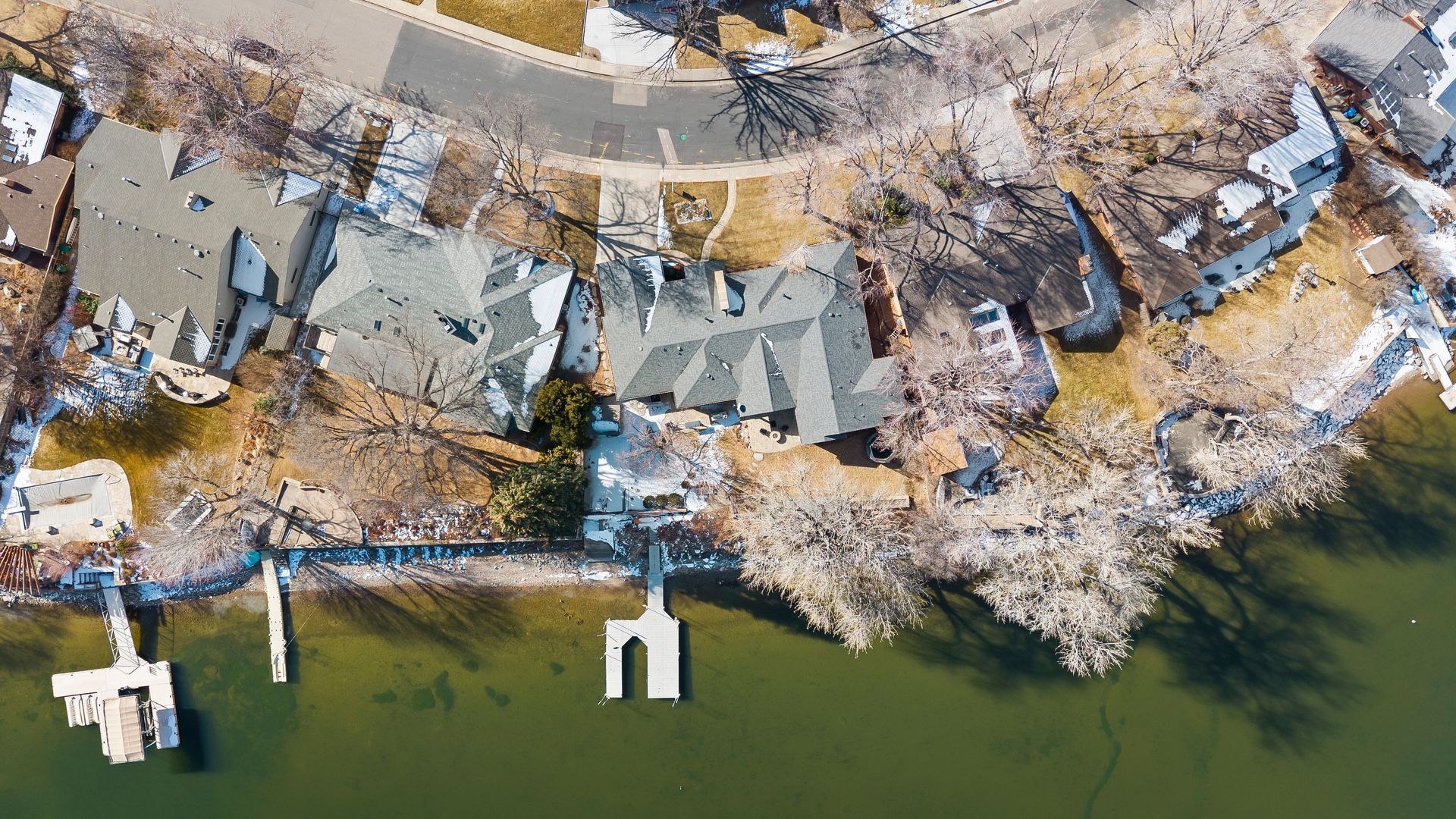 Aerial view of lakefront homes, docks, and a winding road. Snow patches on the ground, trees bare.