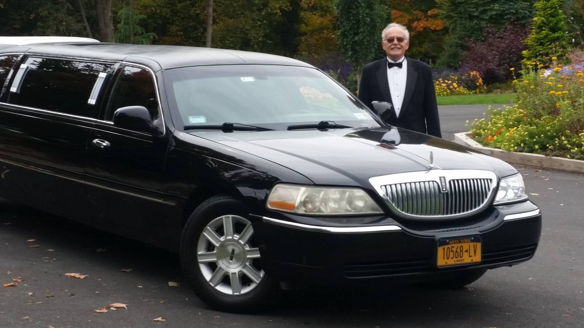 Man in tuxedo stands next to a black limousine outdoors.