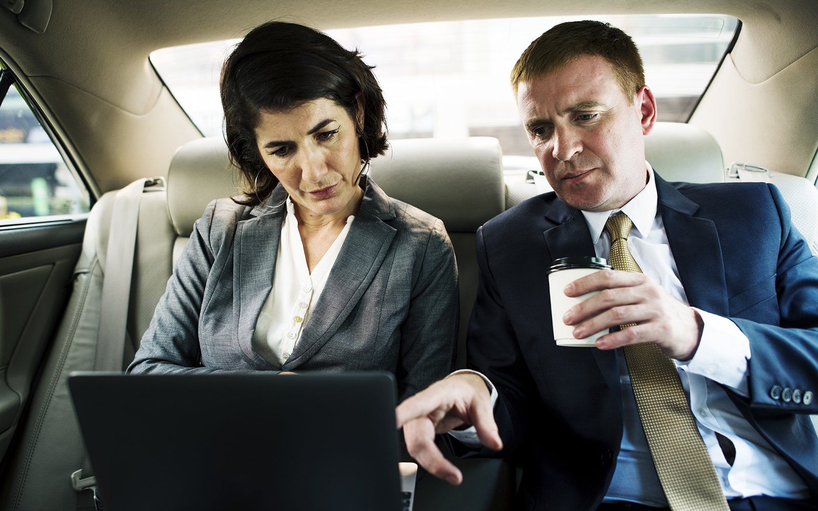 Two businesspeople working on a laptop in a car. Woman looks at screen, man gestures, holding coffee.