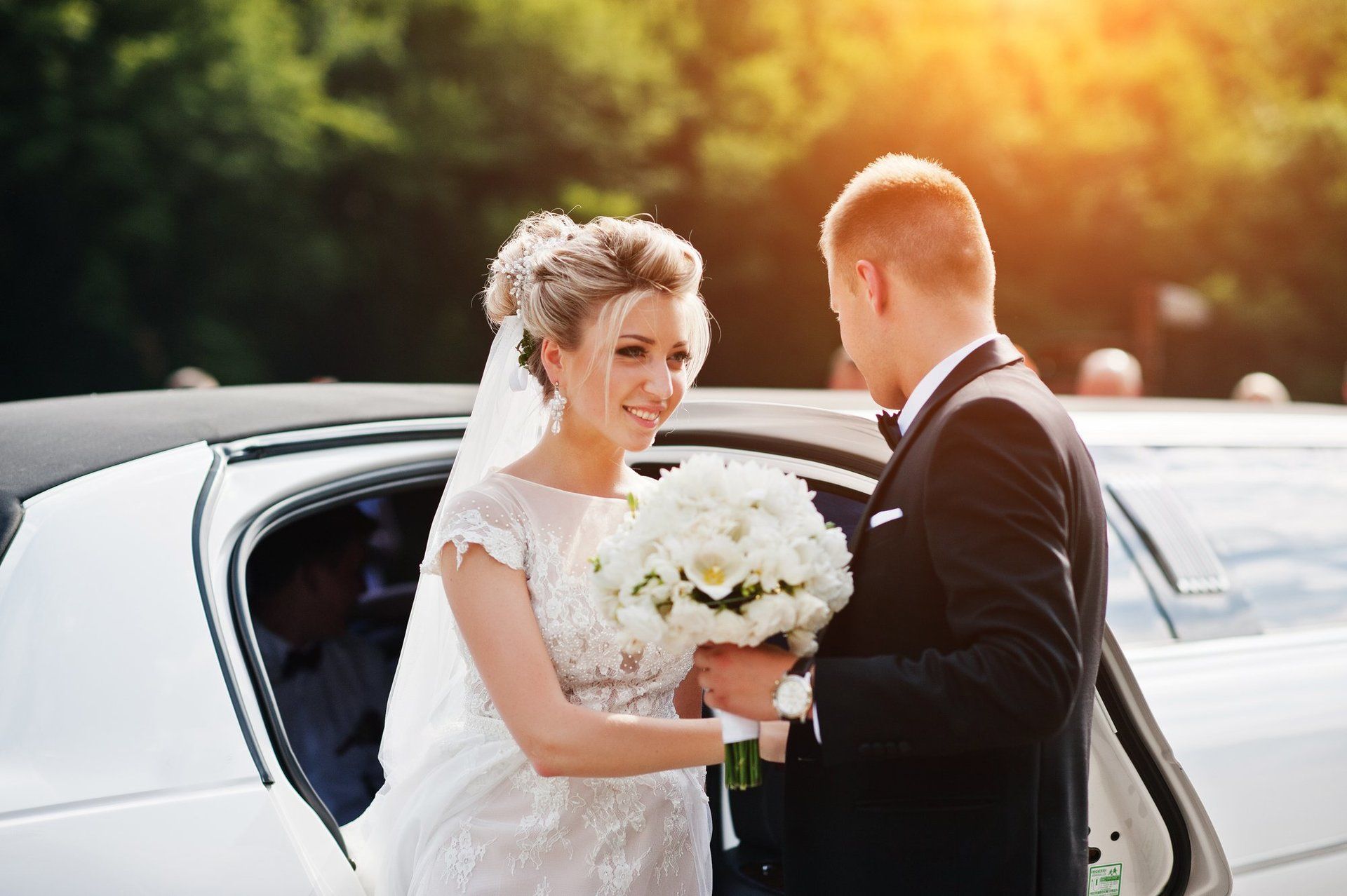Bride and groom smiling near a white limousine, holding a bouquet, sunny outdoor setting.