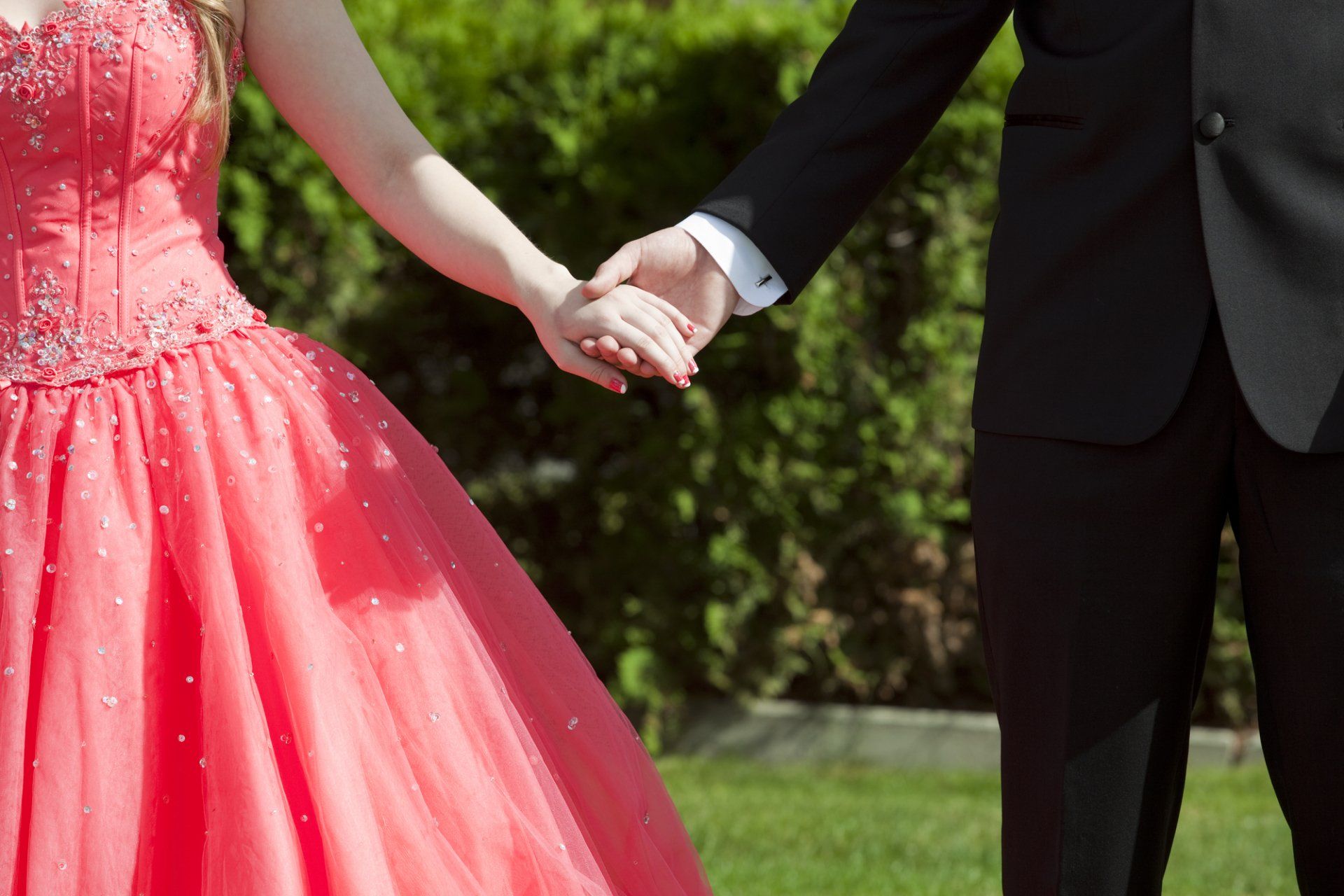 Couple holding hands; woman in coral ball gown, man in black suit, outdoors.
