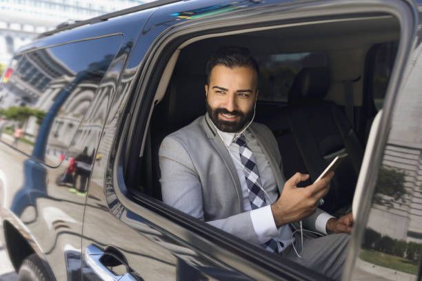 Businessman smiles from the inside of a limousine as he uses a smartphone and earbuds connected to it.