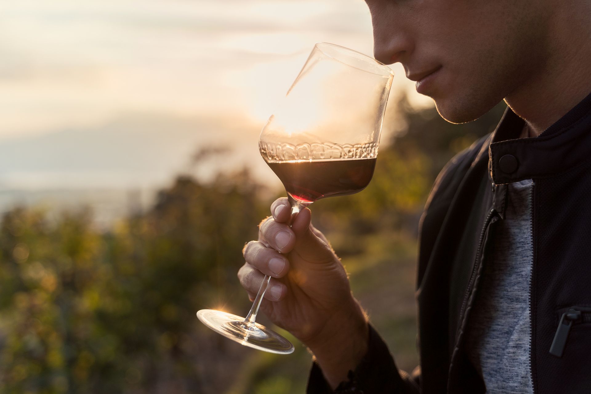 Close-up of a young man as he tastes red wine from a cup, with the sunset in the background.