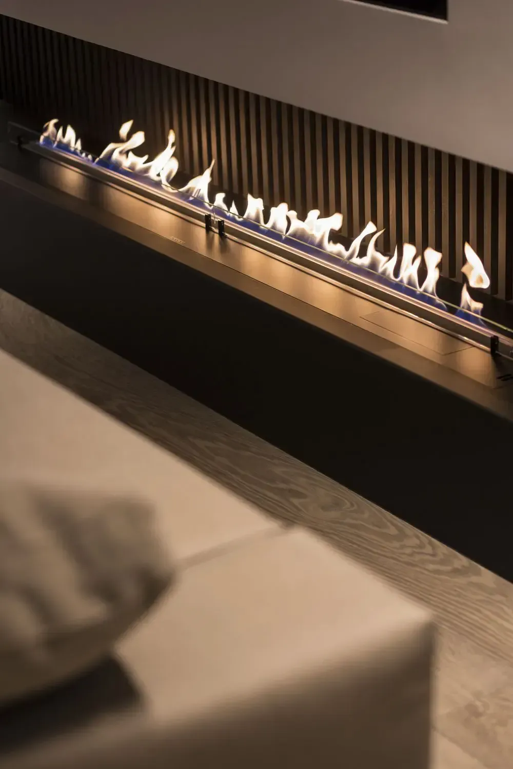 Lungo camino moderno con fiamme contro una parete a doghe, con un divano sfocato in primo piano.