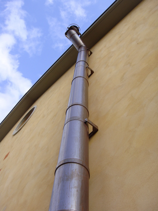 Un tubo di scarico metallico alto e cilindrico, fissato a un edificio color marrone chiaro sotto un cielo nuvoloso.