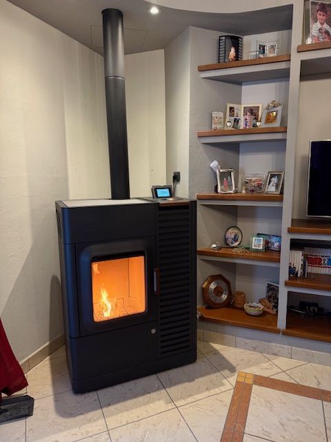 Stufa a pellet nera con fuoco acceso, camino nero. Parete beige con mensole e decorazioni in legno.