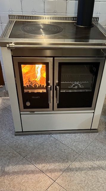 Una stufa a legna con fiamme visibili nel focolare, sportello del forno a destra e piano cottura nero.