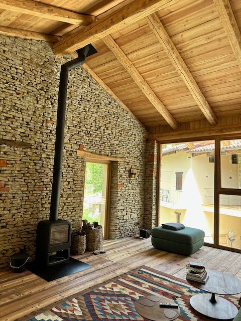 Camera accogliente con stufa a legna, parete in pietra, soffitto in legno e tappeto; ampia finestra con vista.
