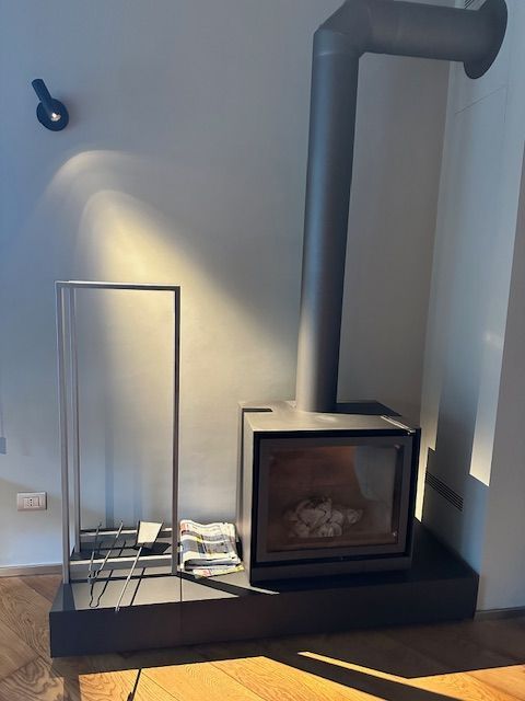 Un moderno camino grigio scuro con una canna fumaria alta, su una piattaforma scura, accanto a un portalegna argentato.