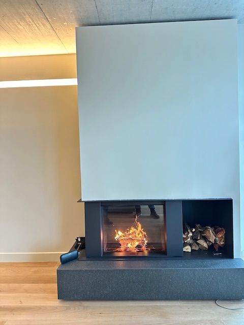Camino moderno con fiamme ardenti, legnaia e una grande parete bianca in una stanza piena di luce.