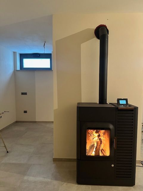 Una stufa a pellet nera con il fuoco acceso in una stanza dalle pareti beige e una piccola finestra.