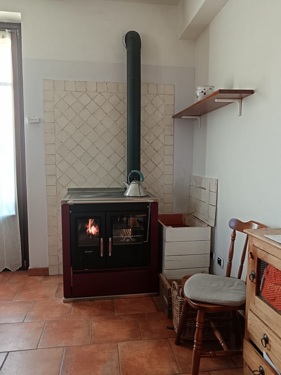 Stufa a legna rossa con tubo nero, paraschizzi piastrellato e una mensola in una stanza con pavimenti piastrellati rossi.