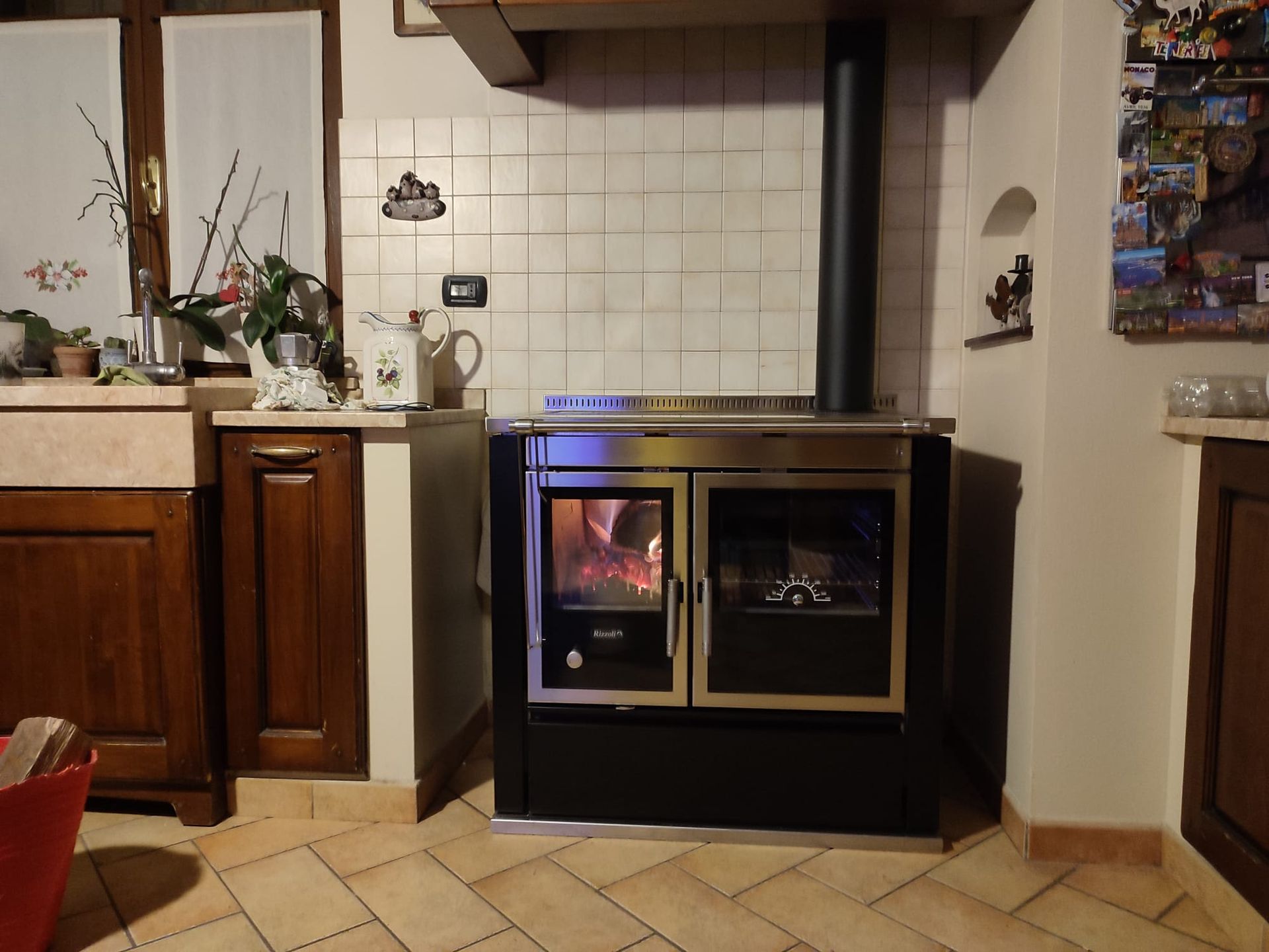 Una stufa a legna con fuoco acceso, in una cucina con mobili scuri e paraschizzi piastrellato.