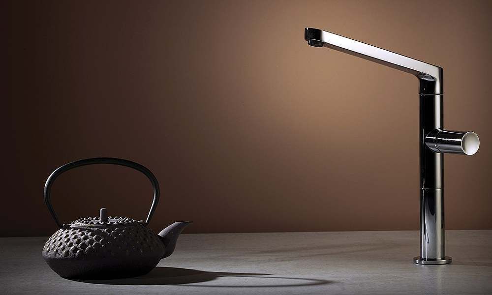 Rubinetto cromato inclinato sopra una teiera su un tavolo, su uno sfondo marrone.