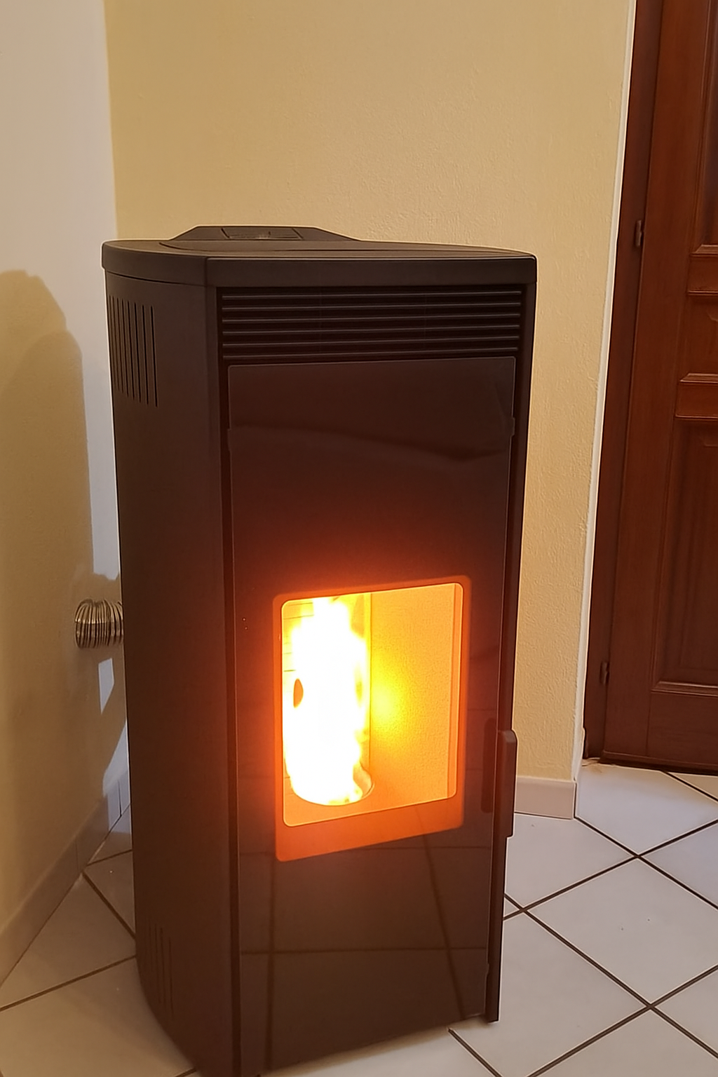 Una stufa a pellet marrone scuro con una fiamma arancione brillante in una stanza, accanto a un muro e a una porta.