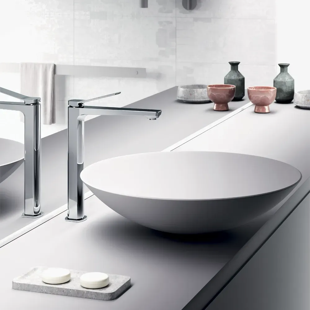 Bagno con lavabo bianco, rubinetti cromati e bottiglie e ciotole decorative su un piano di lavoro grigio chiaro.
