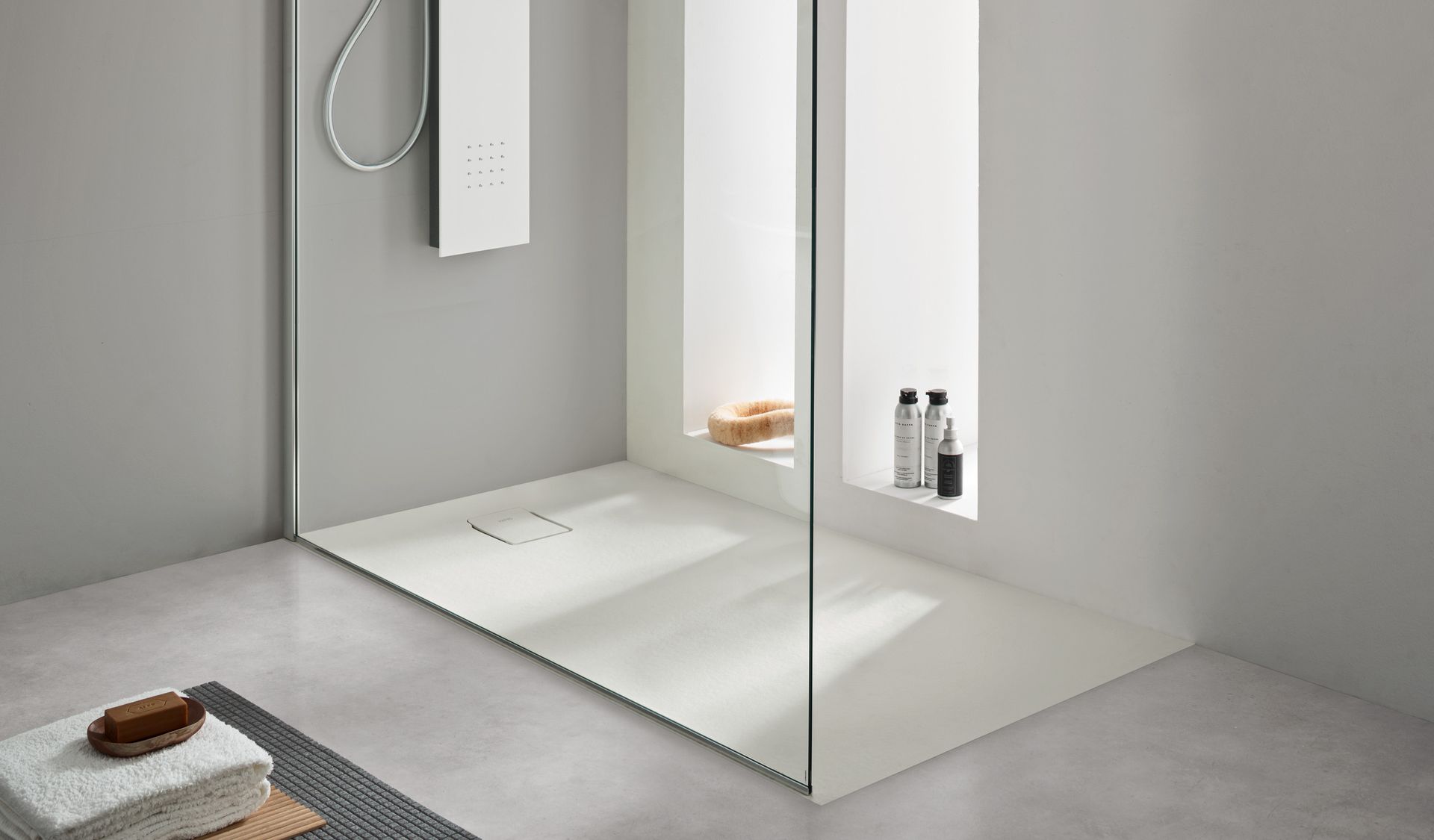 Moderna cabina doccia bianca con pannello in vetro. Pavimento in cemento, pareti grigie, articoli da toeletta su mensola.