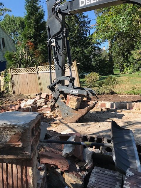 An excavator with a grapple arm is demolishing a brick structure in a residential setting.