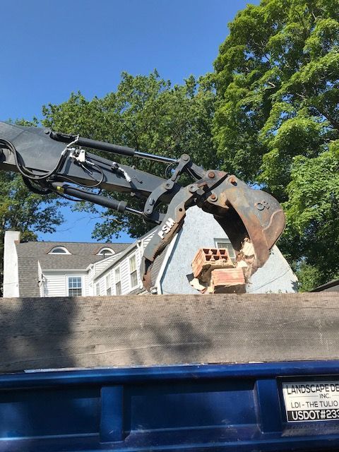 Excavator removing debris from a roof. The machine has a claw, blue sky, and a house in the background.