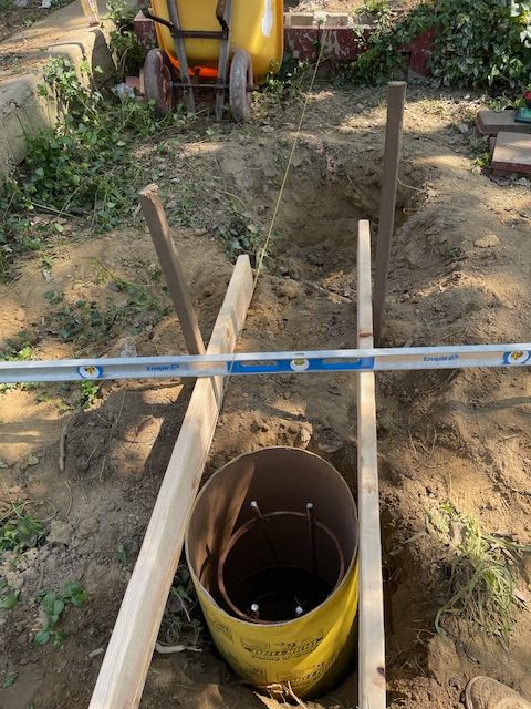 Construction site: A cylindrical form in a hole, wood supports, level, string, wheelbarrow in background.