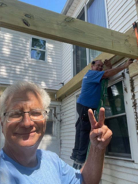 Two men building an outdoor structure; one on a ladder, the other taking a selfie and giving a peace sign.