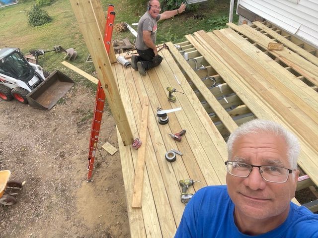 Construction workers building a wooden deck. One man takes a selfie while another works in the background.