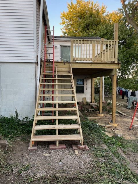 New wooden deck with stairs attached to a white building; ladder leans against stairs; exterior daytime.