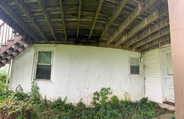Exterior view of a white stucco building with two windows and a wooden deck overhead; green foliage grows around the base.