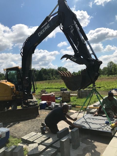 John Deere excavator lifting large stone slab, two men working with blocks outdoors.