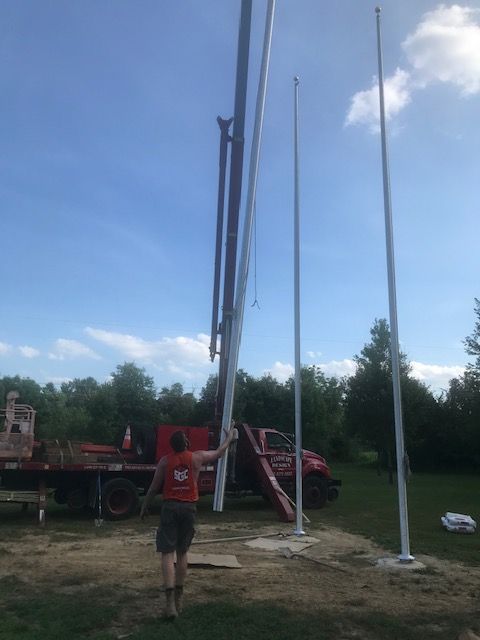Person guiding a long, white pole towards a concrete base. Other poles and machinery visible outdoors.