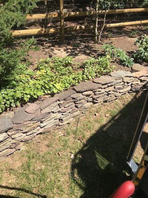 Stone retaining wall bordering a garden bed with lush green plants. Sunlight casts shadows on the grass.
