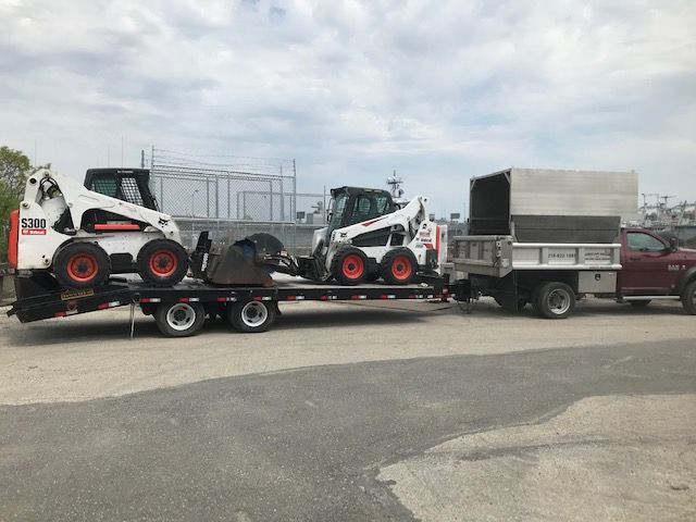 Two Bobcat skid steer loaders on a flatbed trailer pulled by a truck in an industrial setting.