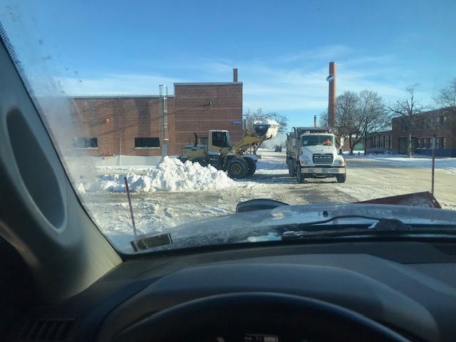 Snow removal equipment clearing a parking lot. A front-end loader and a plow truck work together on a sunny day.