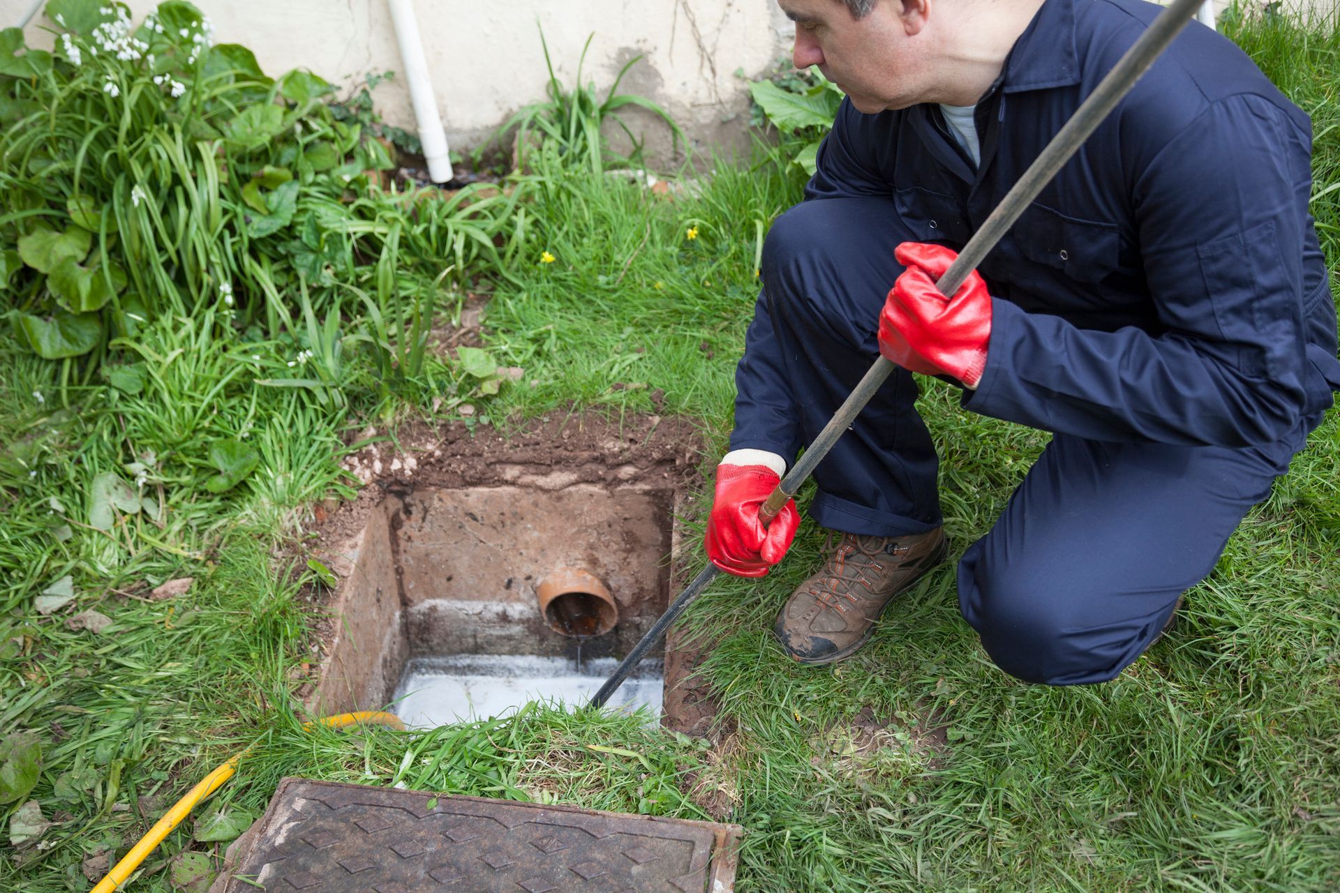 Plumber in blue uniform and red gloves cleaning outdoor drain with rod in grassy yard. Plumber in blue uniform and red gloves cleaning outdoor drain with rod in grassy yard.