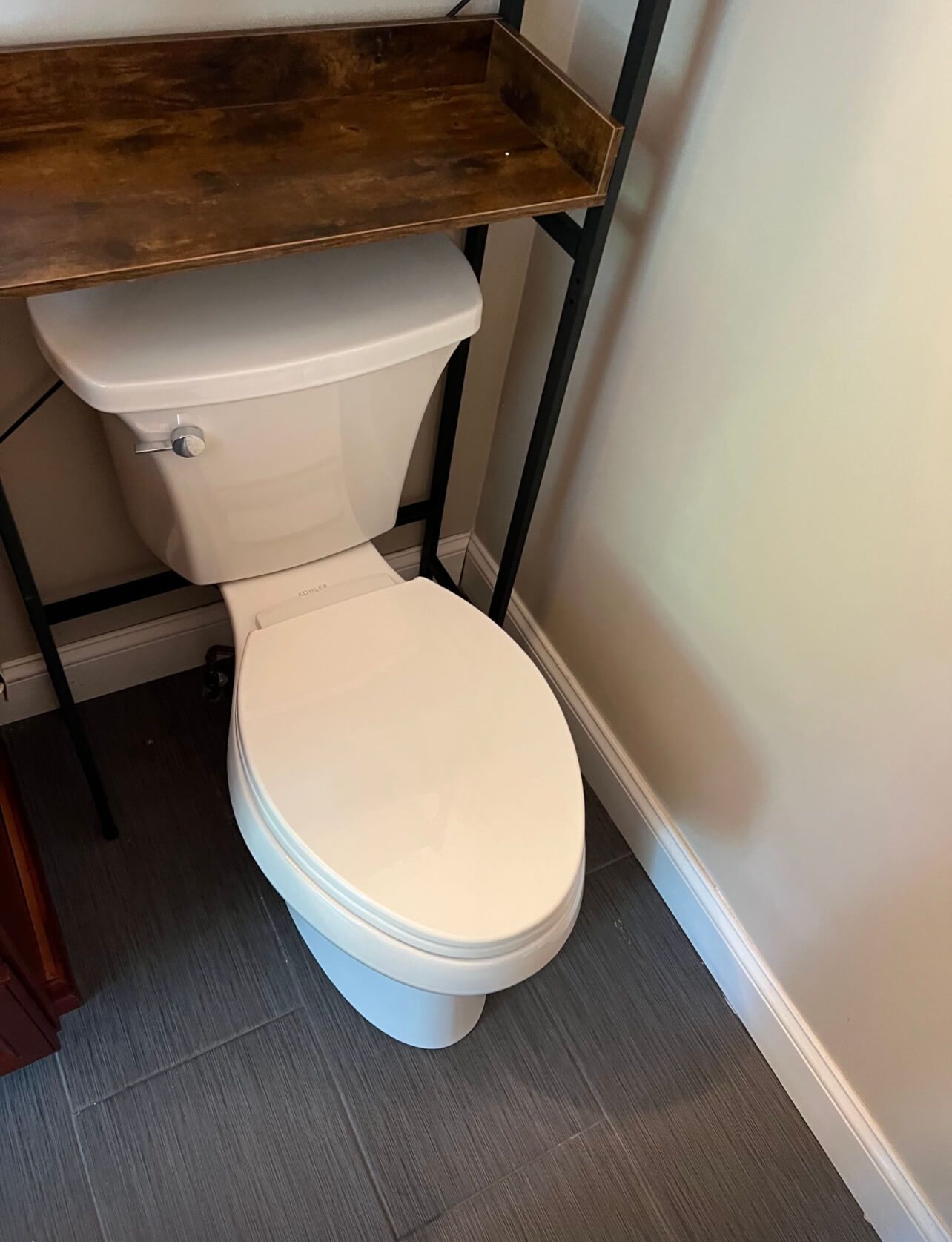 A toilet is sitting under a wooden shelf in a bathroom.