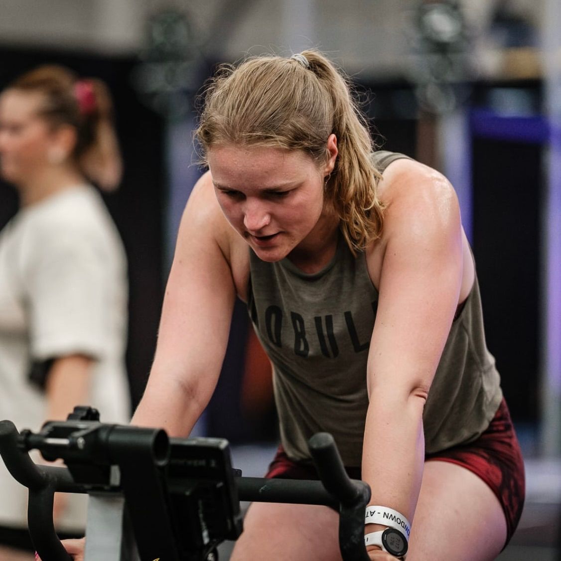 Vrouw in een tanktop op een hometrainer, naar beneden kijkend, zwetend in een sportschoolomgeving.
