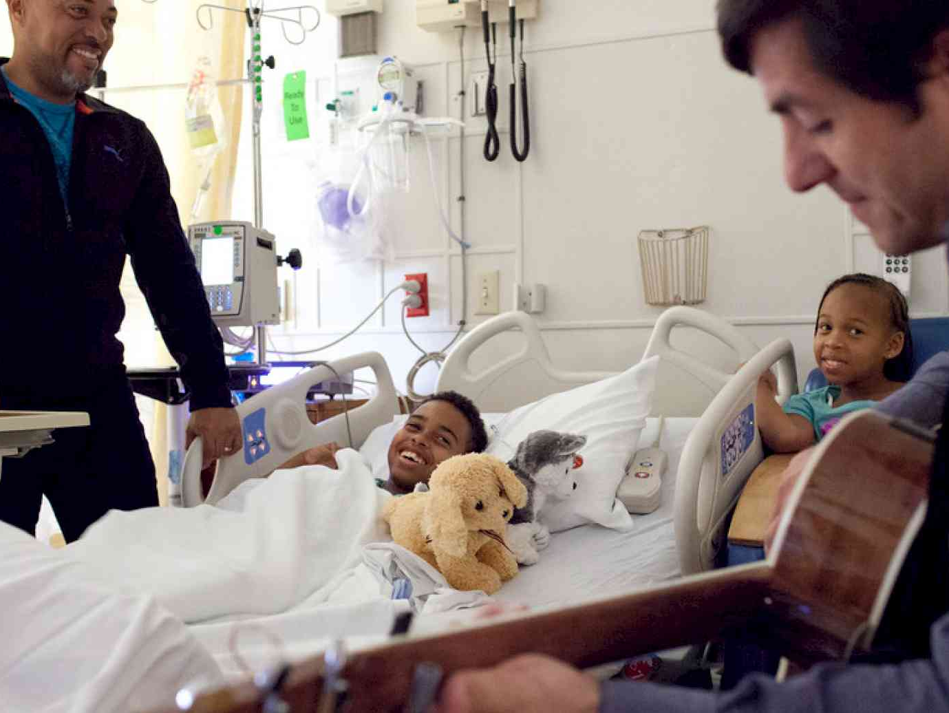 Boy laying in hospital bed looking up with a smile at a musician playing the guitar at his bedside. His father is smiling across from the musician on the other side of the bed.
