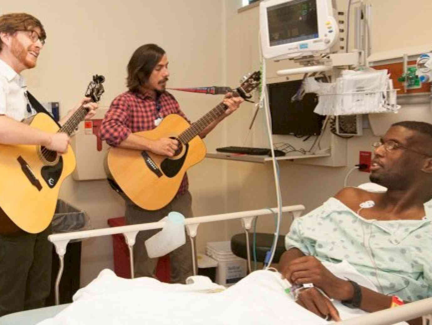 Two guitar players playing at the bedside of a patient laying in a hospital bed.