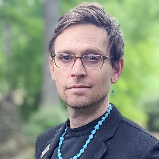 A man wearing glasses and a blue necklace is looking at the camera.