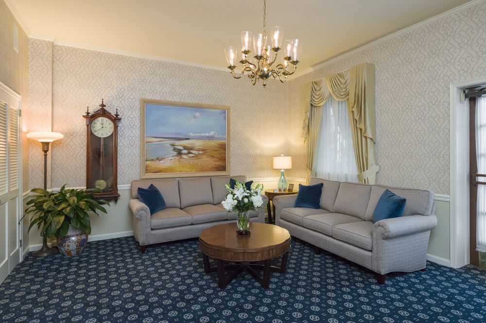 Formal visitation room with gray seating, traditional chandelier, grandfather clock, and scenic artwork