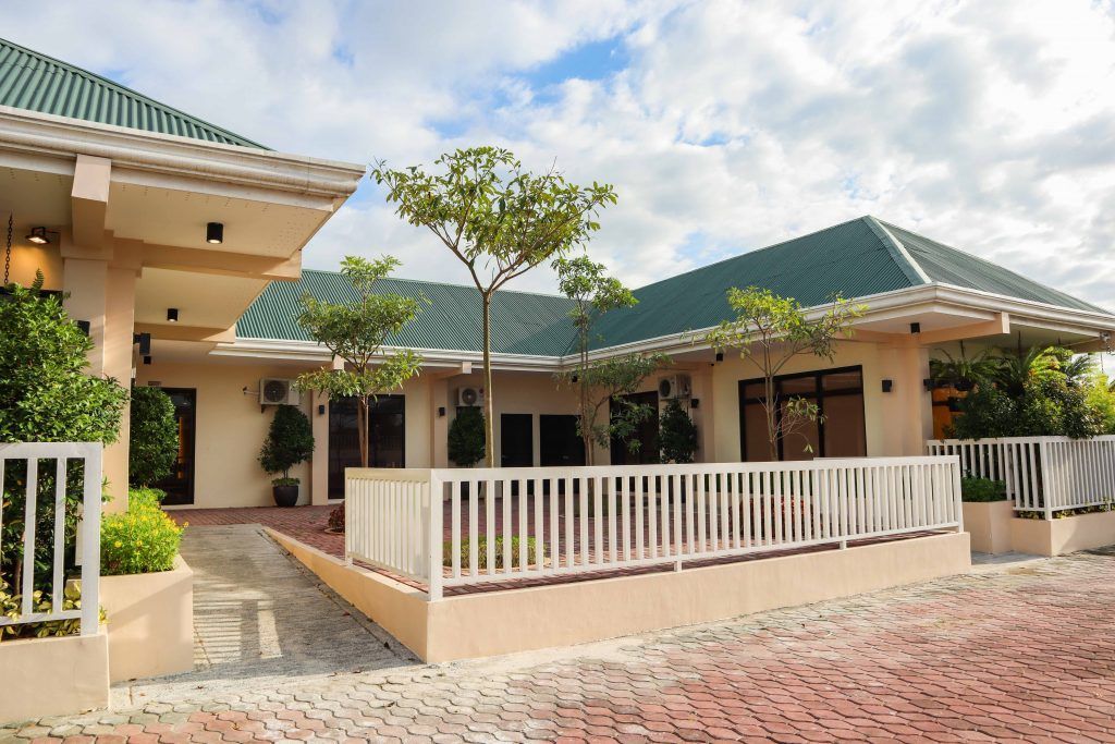 Single-story funeral home with green metal roof, white fencing, covered walkways, and tropical landscaping