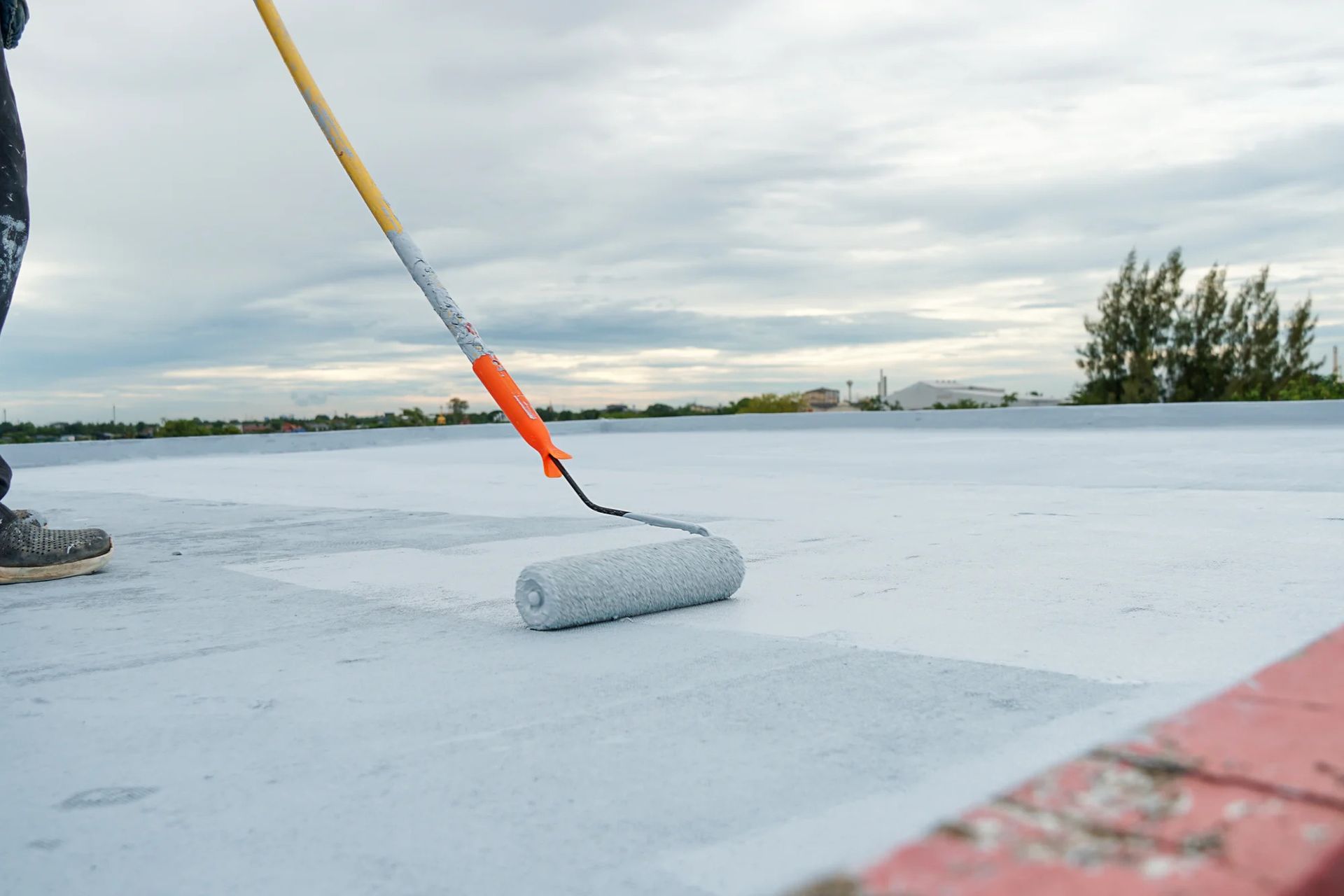 Worker applying white acrylic roof coating with roller on commercial flat roof surface.