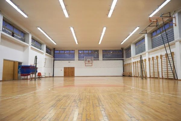 School gymnasium with wooden basketball court, fluorescent lighting, wall bars, equipment storage, and high windows