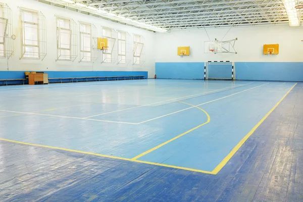Indoor gymnasium with blue floor markings, basketball hoops, soccer goals, and exercise equipment storage area
