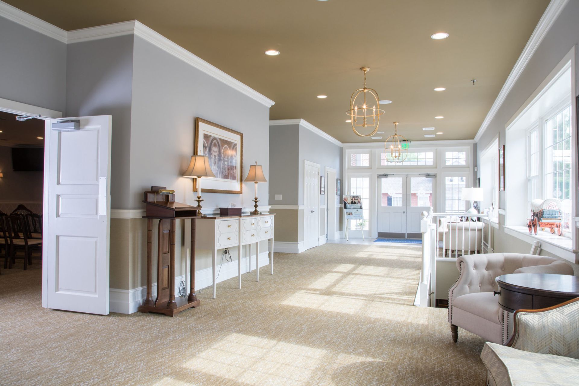 Elegant funeral home lobby with gray walls, traditional furnishings, brass lighting fixtures, and large windows