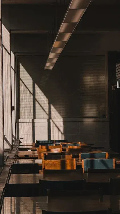 Sunlit wooden classroom with traditional desks, exposed beam ceiling, and large windows creating dramatic shadows